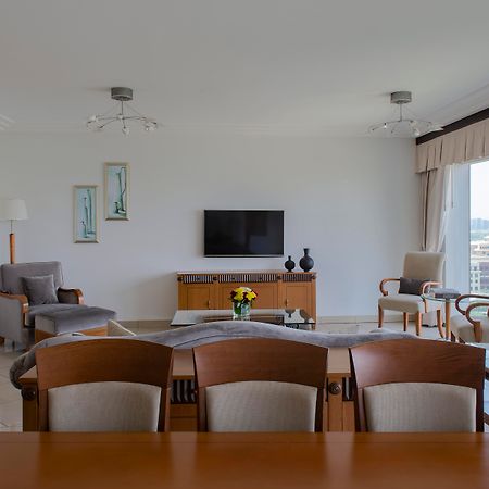 Grand Hyatt Dubai Hotel Exterior photo The photo shows a modern living room with a spacious and minimalistic design. There is a flat-screen television mounted on the wall, facing a seating arrangement that includes two armchairs and a sofa. A wooden table is positioned in the foreground, 