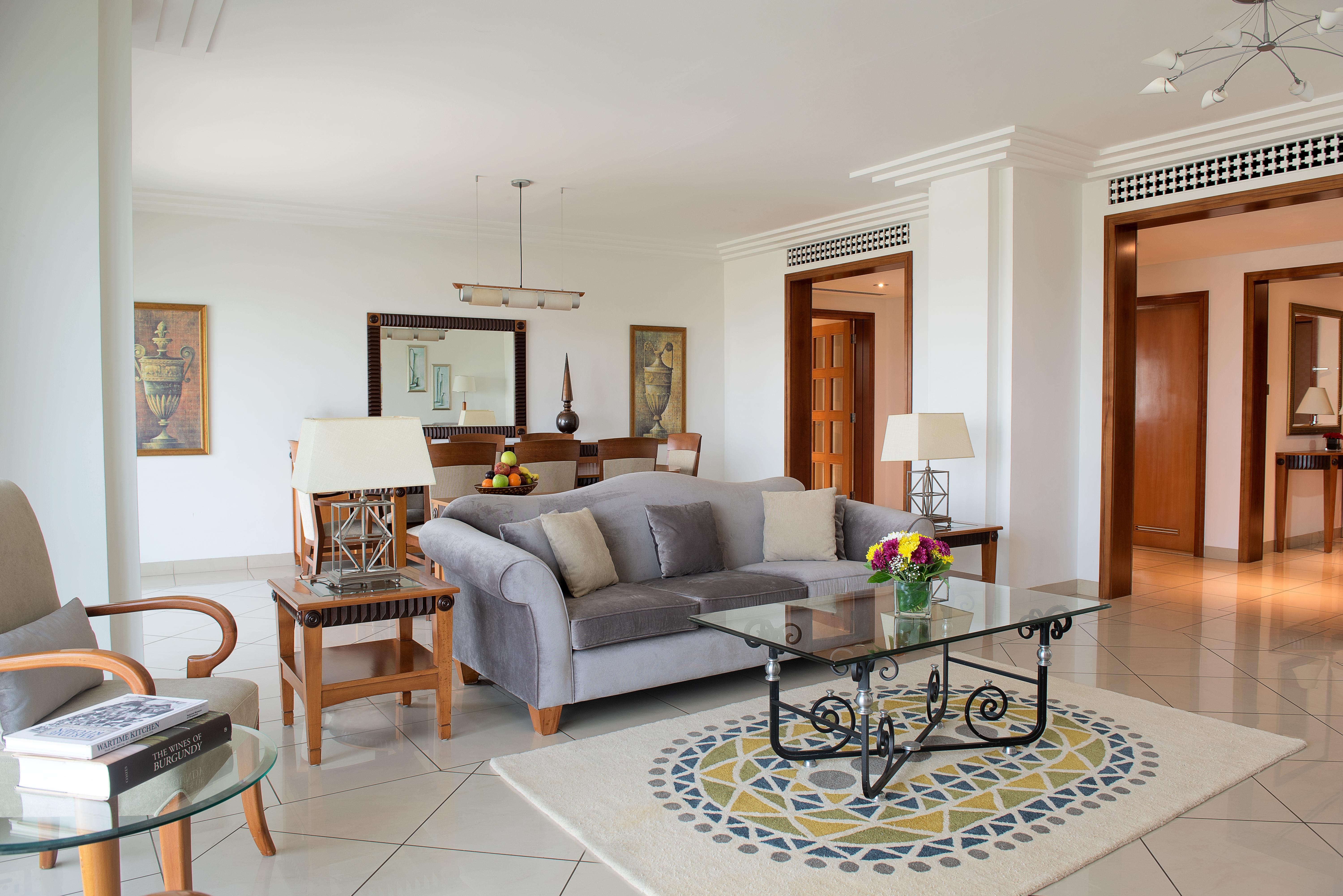 Grand Hyatt Dubai Hotel Exterior photo The photo shows a spacious and well-lit living room area. It features a light gray sofa with plush cushions, positioned around a glass coffee table with an ornate black metal base. A circular area rug with a decorative pattern is placed beneath the t