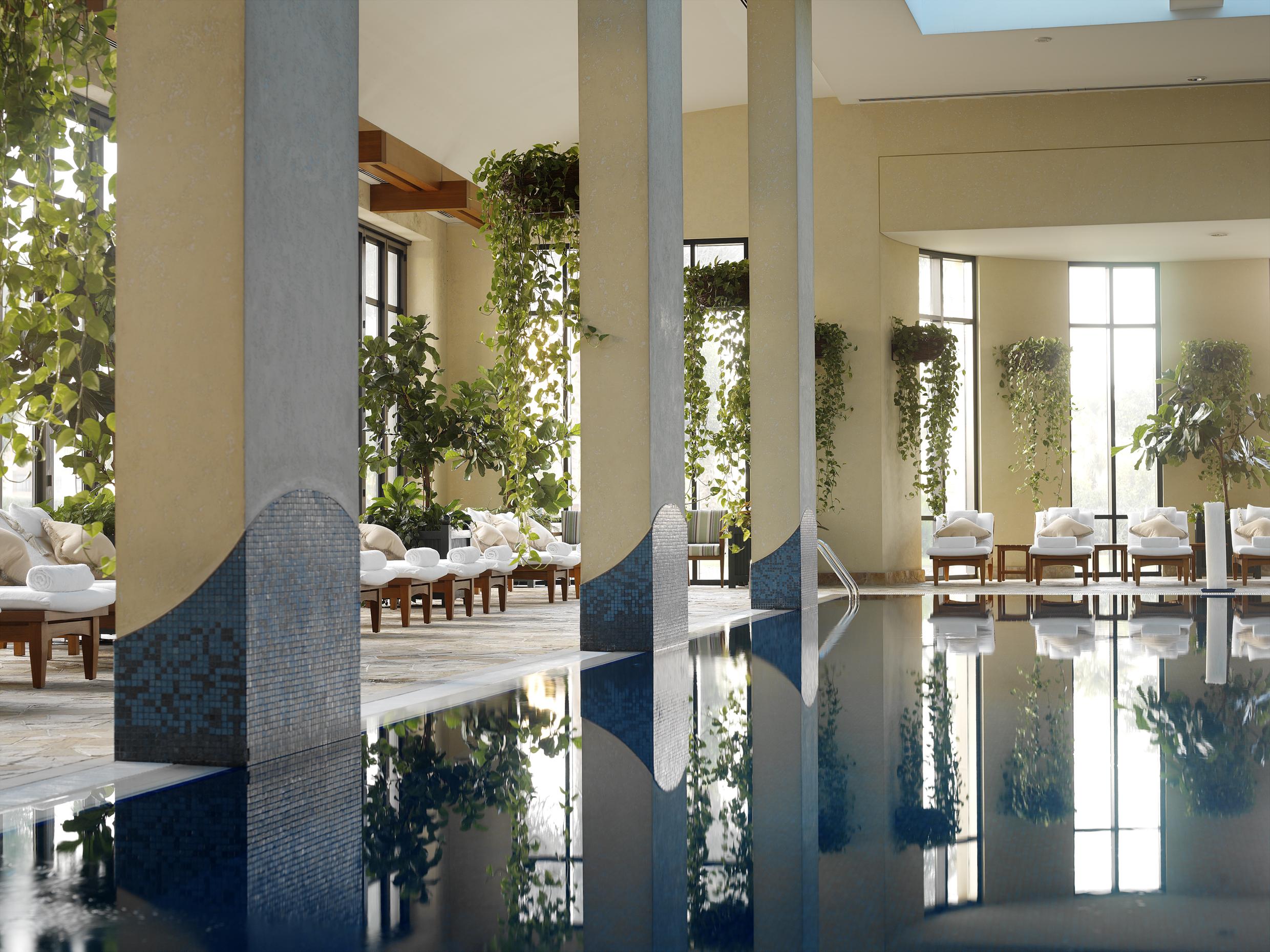 Grand Hyatt Dubai Hotel Facilities photo The image depicts a serene indoor swimming pool area. It features large windows allowing natural light to stream in, creating a bright and inviting atmosphere. The pool's calm waters reflect the surroundings, including decorative pillars and lush gre