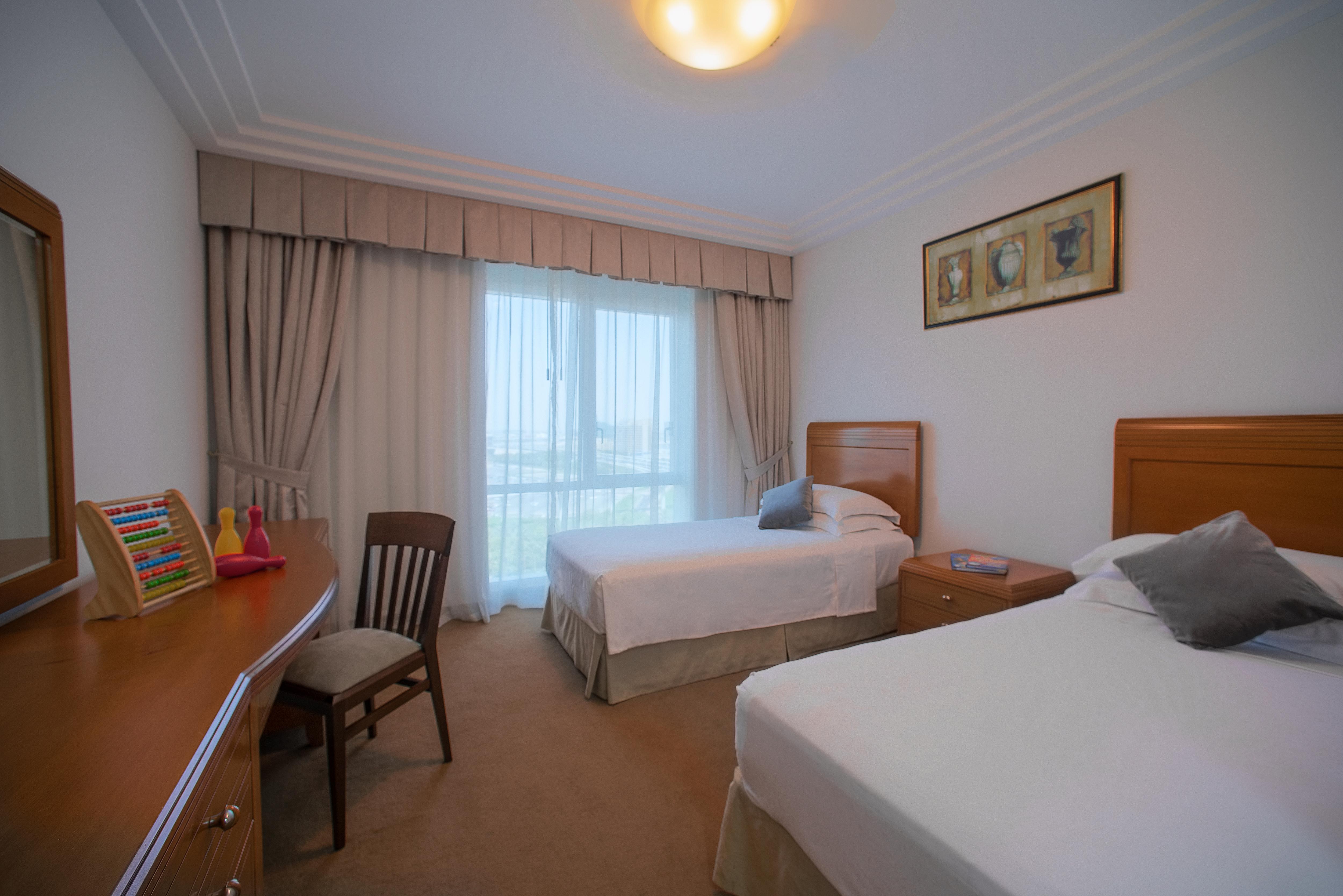 Grand Hyatt Dubai Hotel Exterior photo The photo shows a spacious hotel room featuring two single beds with white bedding, positioned against a wall. There is a wooden dresser next to the bed, along with a chair. A large window, covered with light-colored curtains, allows natural light to