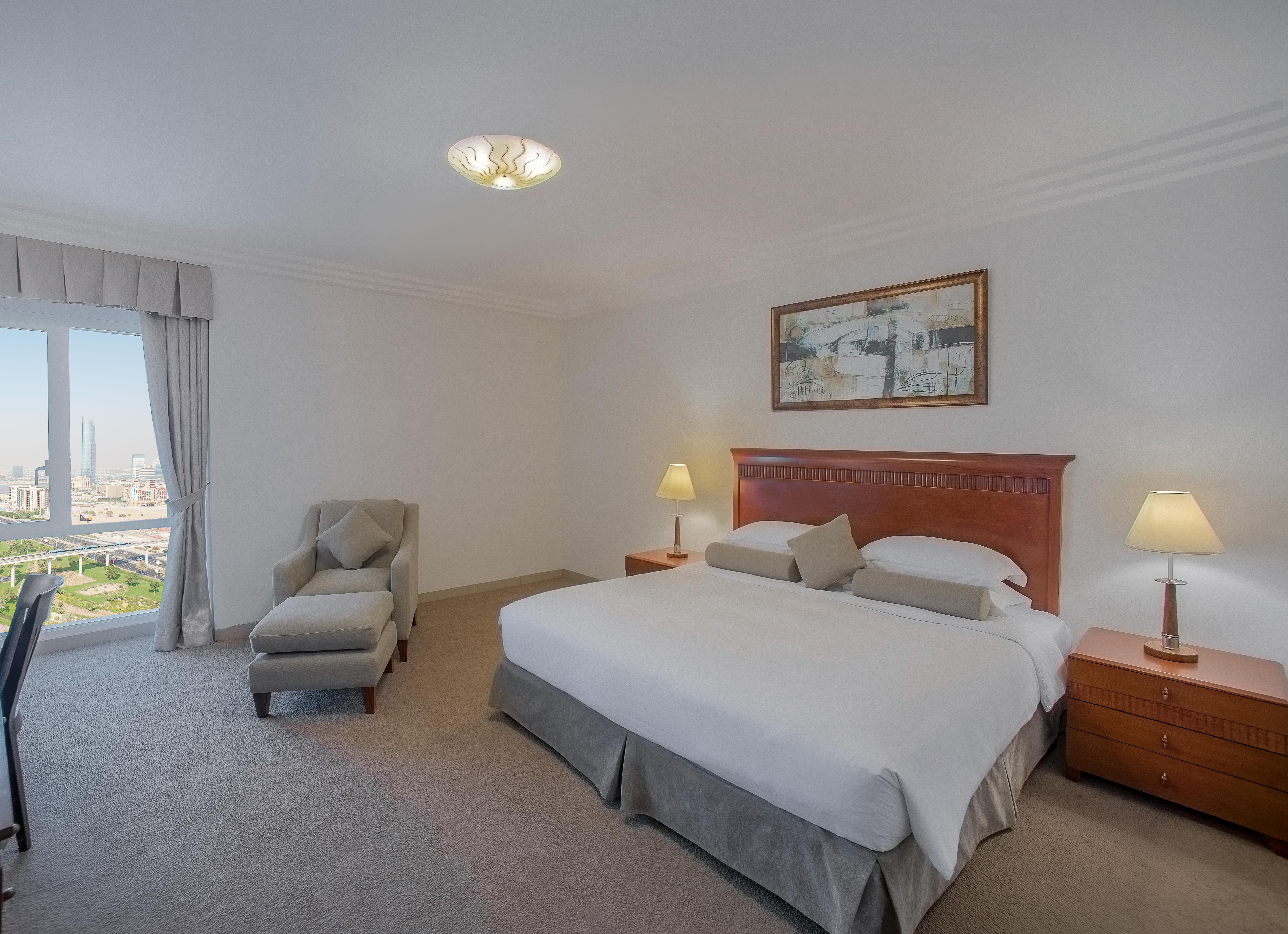 Grand Hyatt Dubai Hotel Exterior photo The photo shows a spacious hotel room with a contemporary design. There is a large bed with white linens and two decorative pillows. Next to the bed are two bedside tables with lamps. On one wall, there is a framed artwork. A comfortable armchair and