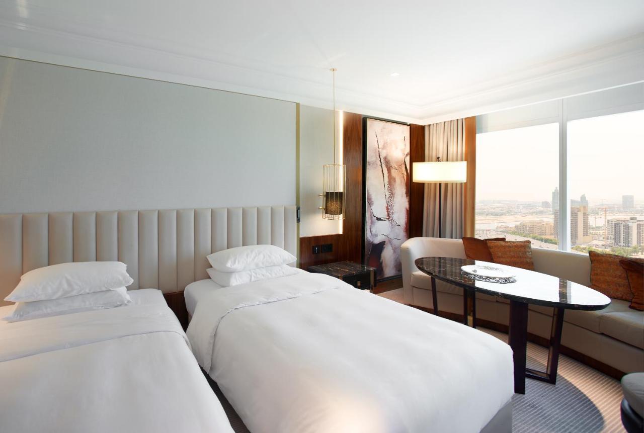 Grand Hyatt Dubai Hotel Exterior photo The photo shows a modern hotel room featuring two separate beds with white bedding. The walls are painted in neutral tones, and there is a large window that allows natural light to enter, offering a view of the city. A stylish round table and chairs 