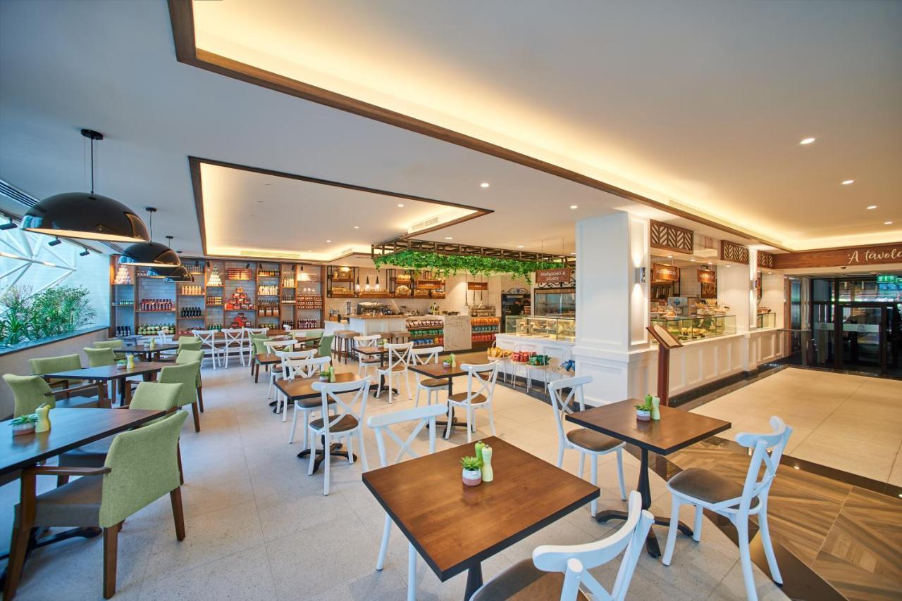 Grand Hyatt Dubai Hotel Exterior photo The photo shows a modern café or restaurant interior. It features a spacious layout with wooden tables and chairs, some of which have white frames and green cushions. The walls are adorned with shelves showcasing various items, and there is a display