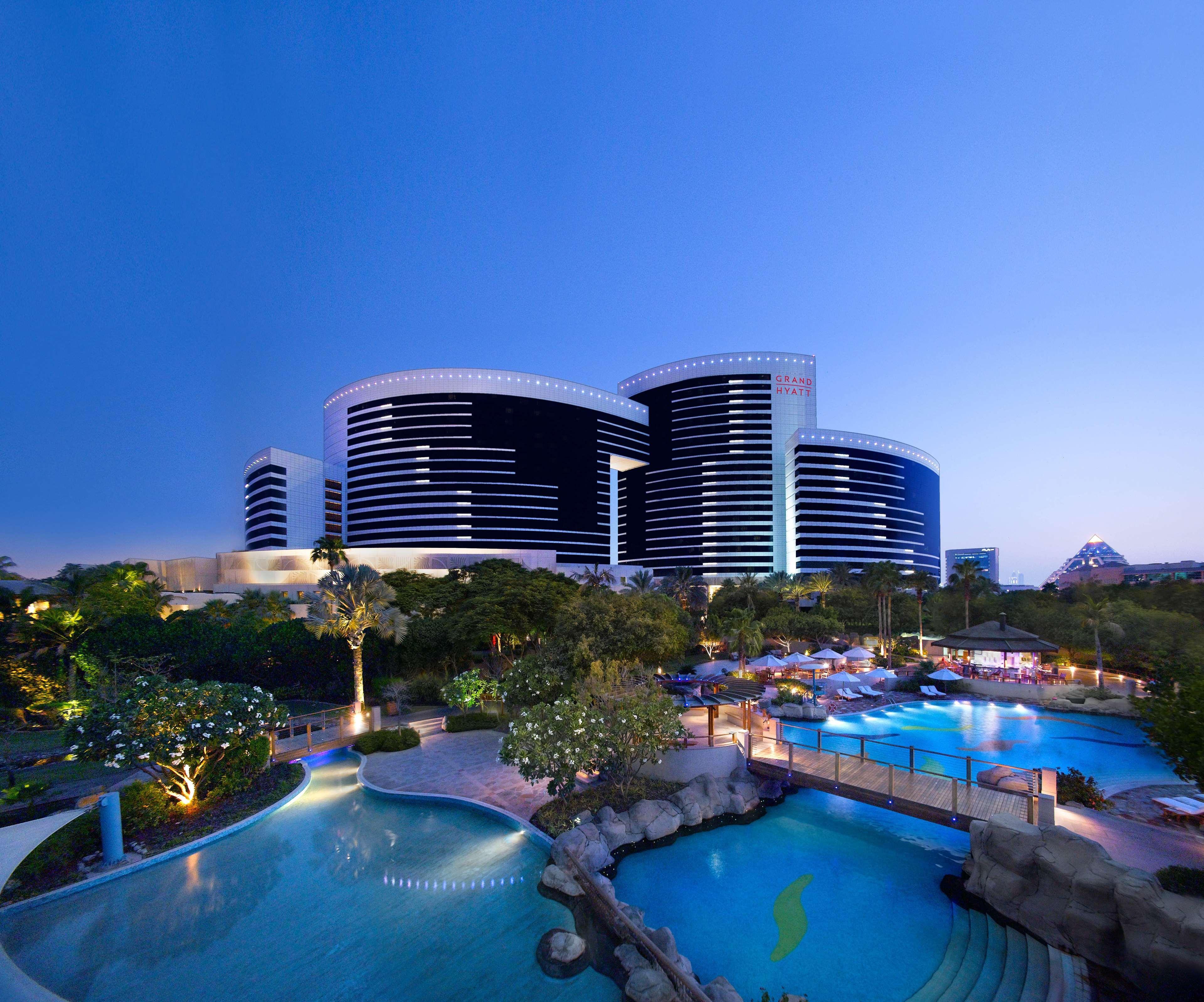 Grand Hyatt Dubai Hotel Exterior photo The photo shows a modern hotel complex featuring several sleek, high-rise buildings with a contemporary architectural design. The buildings have dark facades with wide horizontal bands, giving them a striking appearance against the evening sky. In th