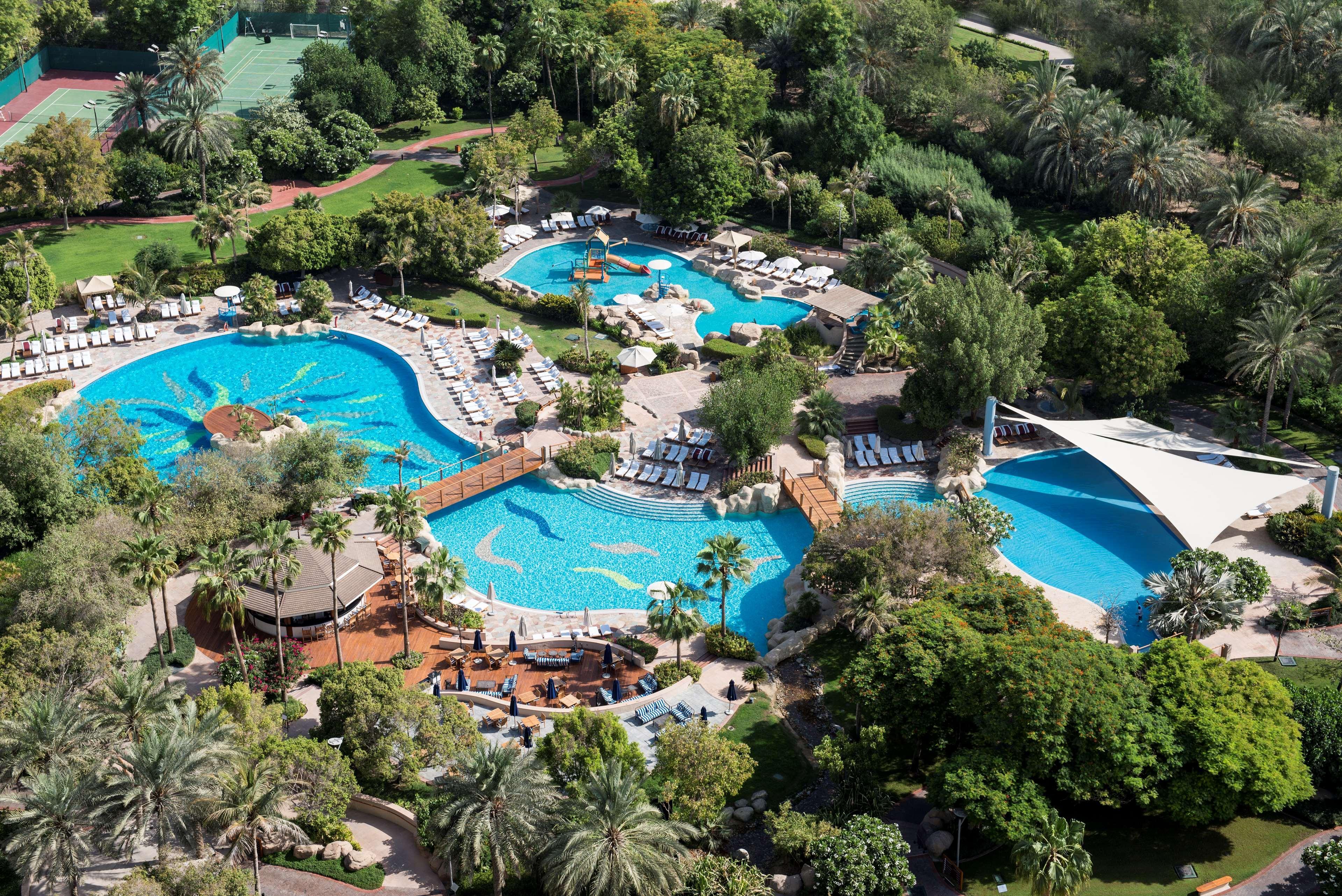 Grand Hyatt Dubai Hotel Exterior photo The photo shows a beautiful aerial view of a luxurious pool area surrounded by lush greenery. There are several interconnected swimming pools of different shapes and sizes, with sun loungers arranged around them. The pools feature various elements su
