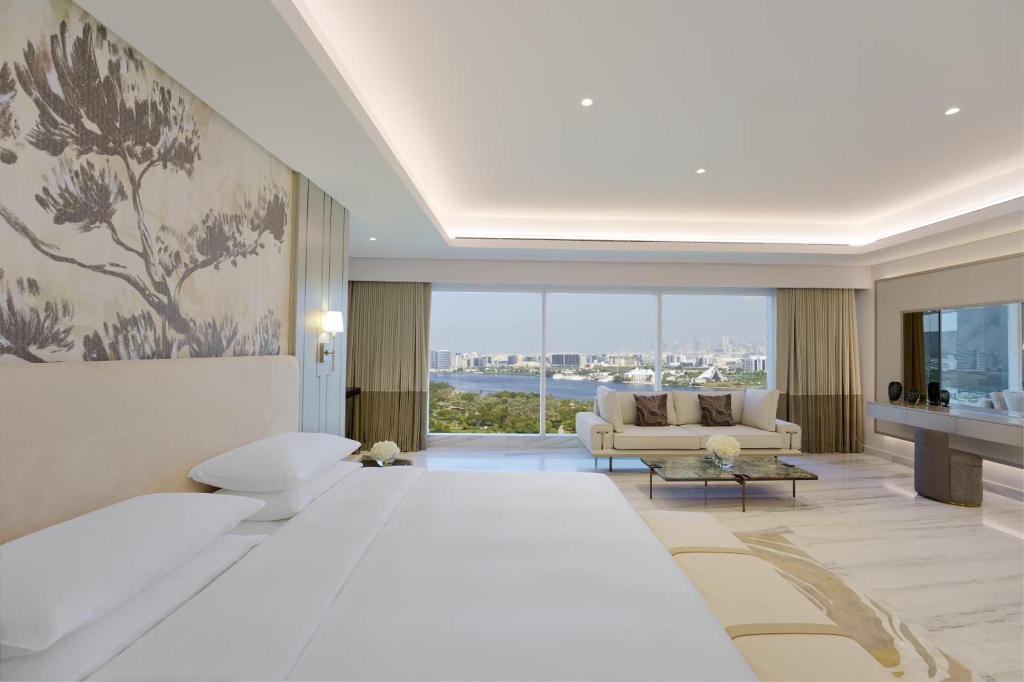 Grand Hyatt Dubai Hotel Exterior photo The photo shows a modern, elegantly designed hotel room. It features a large bed with white linens, positioned against a beautifully decorated wall with a subtle nature-themed mural. There is a cozy seating area with a light-colored sofa and decorati