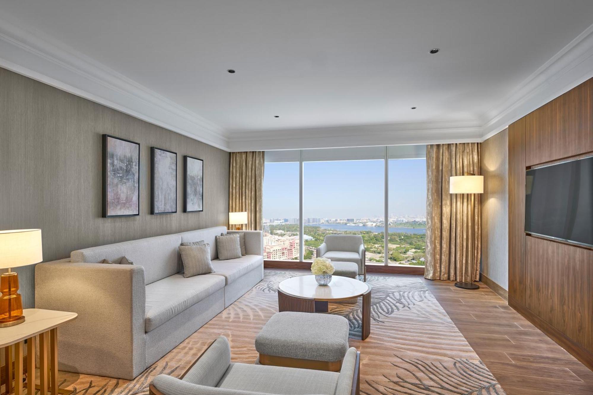 Grand Hyatt Dubai Hotel Exterior photo The photo shows a modern hotel room featuring a comfortable living area. There is a light-colored sofa with several cushions, a coffee table in the center, and a couple of armchairs. The walls are adorned with framed artworks, and large windows provi