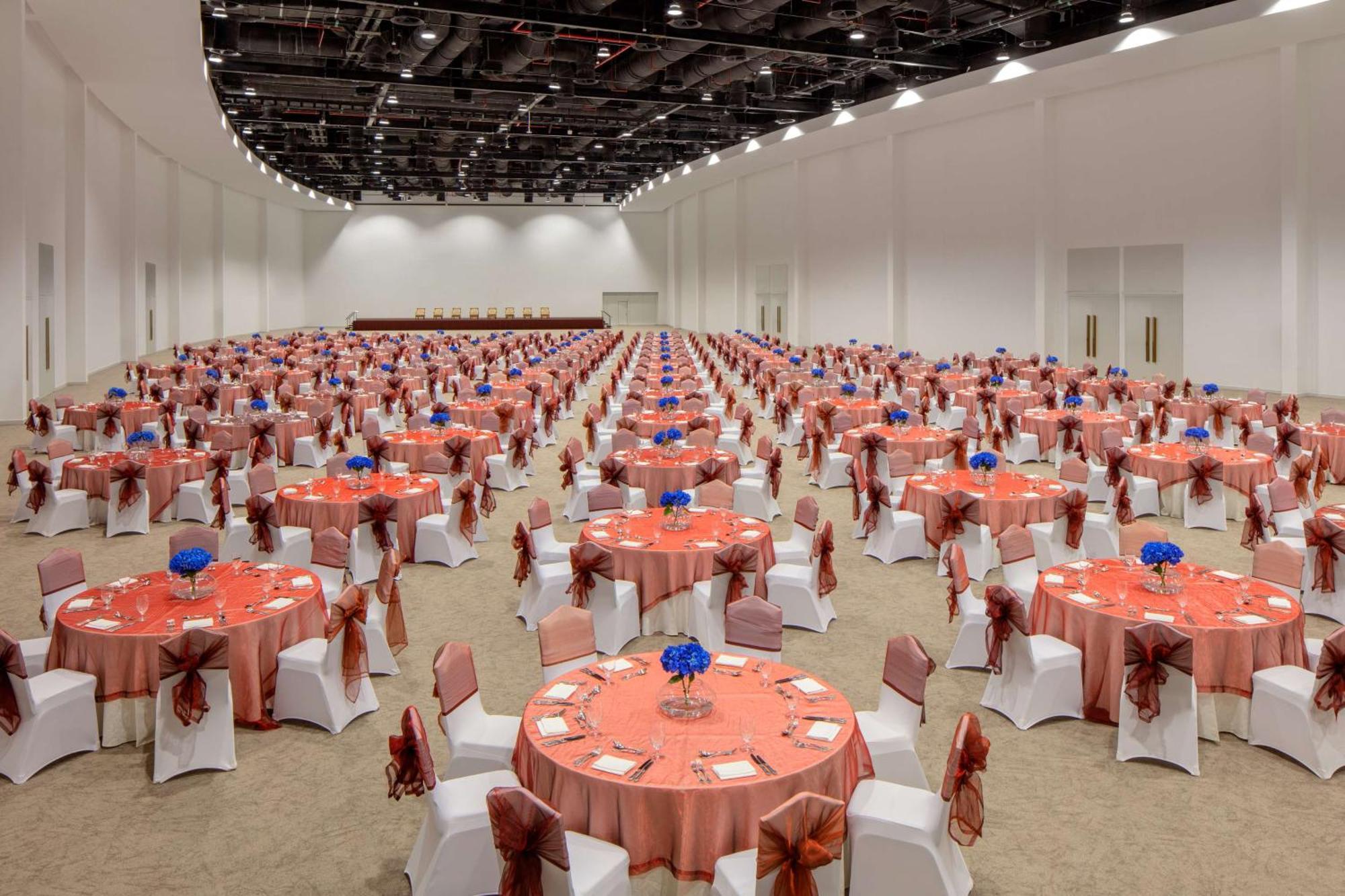 Grand Hyatt Dubai Hotel Exterior photo The photo shows a large banquet hall set up for an event. There are numerous round tables, each covered with orange tablecloths, and decorated with blue flower arrangements in the center. The chairs are white, adorned with orange sashes. The space ap