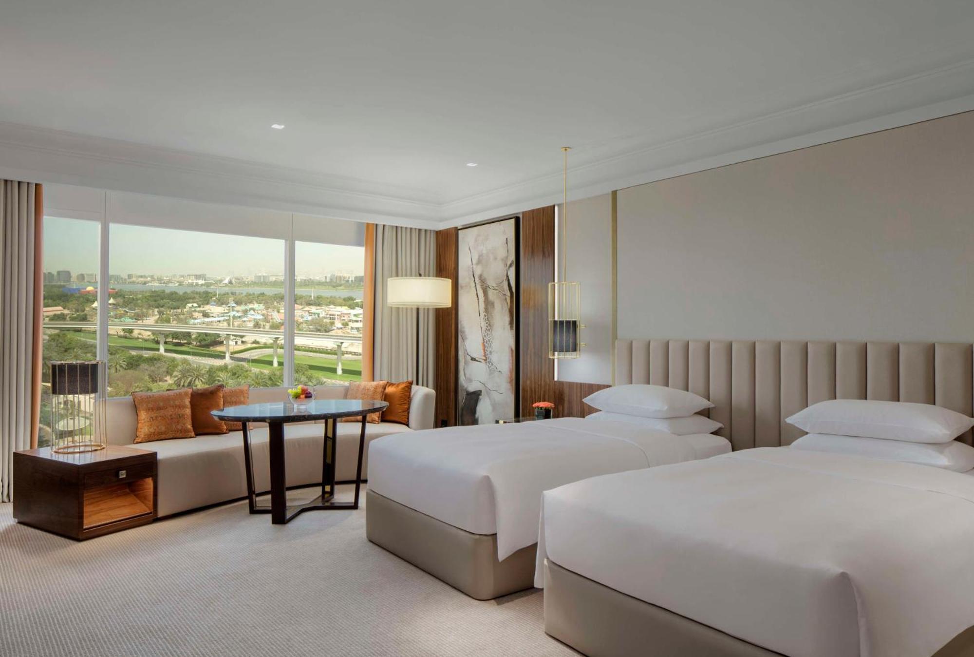 Grand Hyatt Dubai Hotel Exterior photo The photo shows a well-designed hotel room featuring two twin beds with white linens. The room has modern furnishings, including a small round table with chairs, and a large window that offers a view of the outside landscape. Soft lighting and neutra