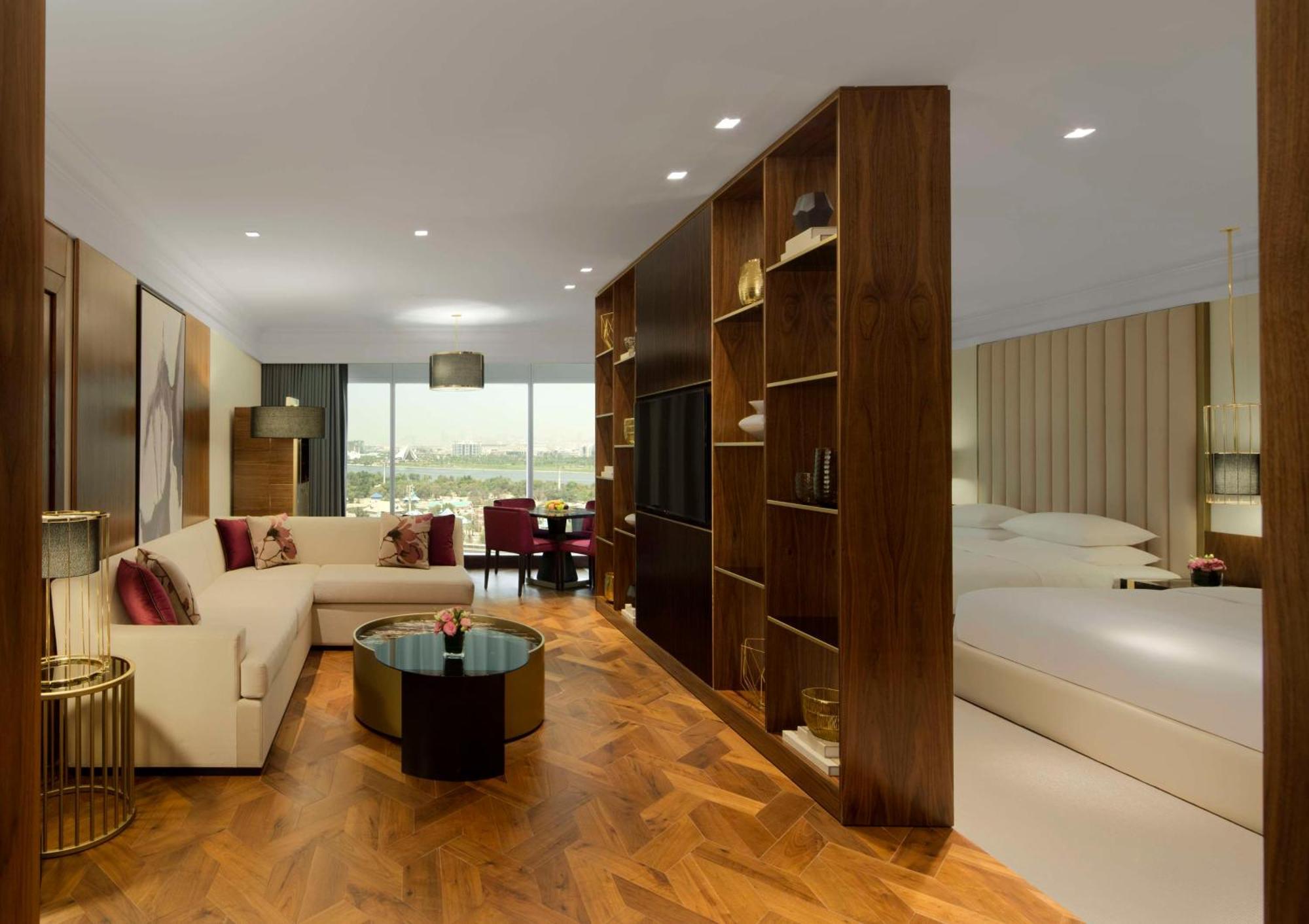 Grand Hyatt Dubai Hotel Exterior photo The photo shows a modern hotel room designed with a stylish and comfortable aesthetic. On one side, there is a cozy seating area featuring a light-colored sofa with decorative cushions and a small coffee table. In the center of the room, a wooden par