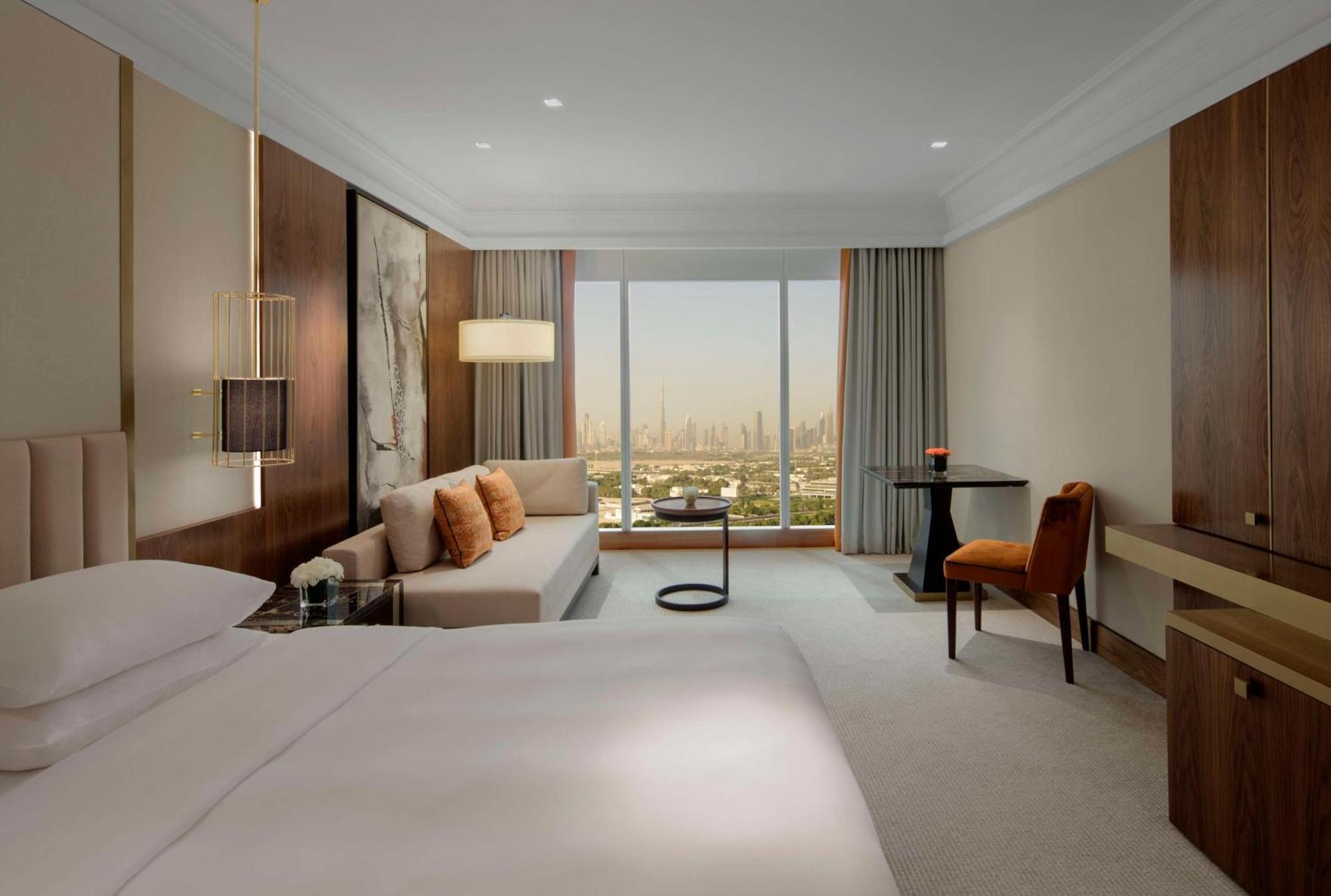 Grand Hyatt Dubai Hotel Exterior photo The photo shows a modern hotel room with a spacious design. It features a large bed with white linens, a comfortable couch adorned with pillows, and a small round table. A stylish chair sits next to a desk or dresser. The room has large windows offer